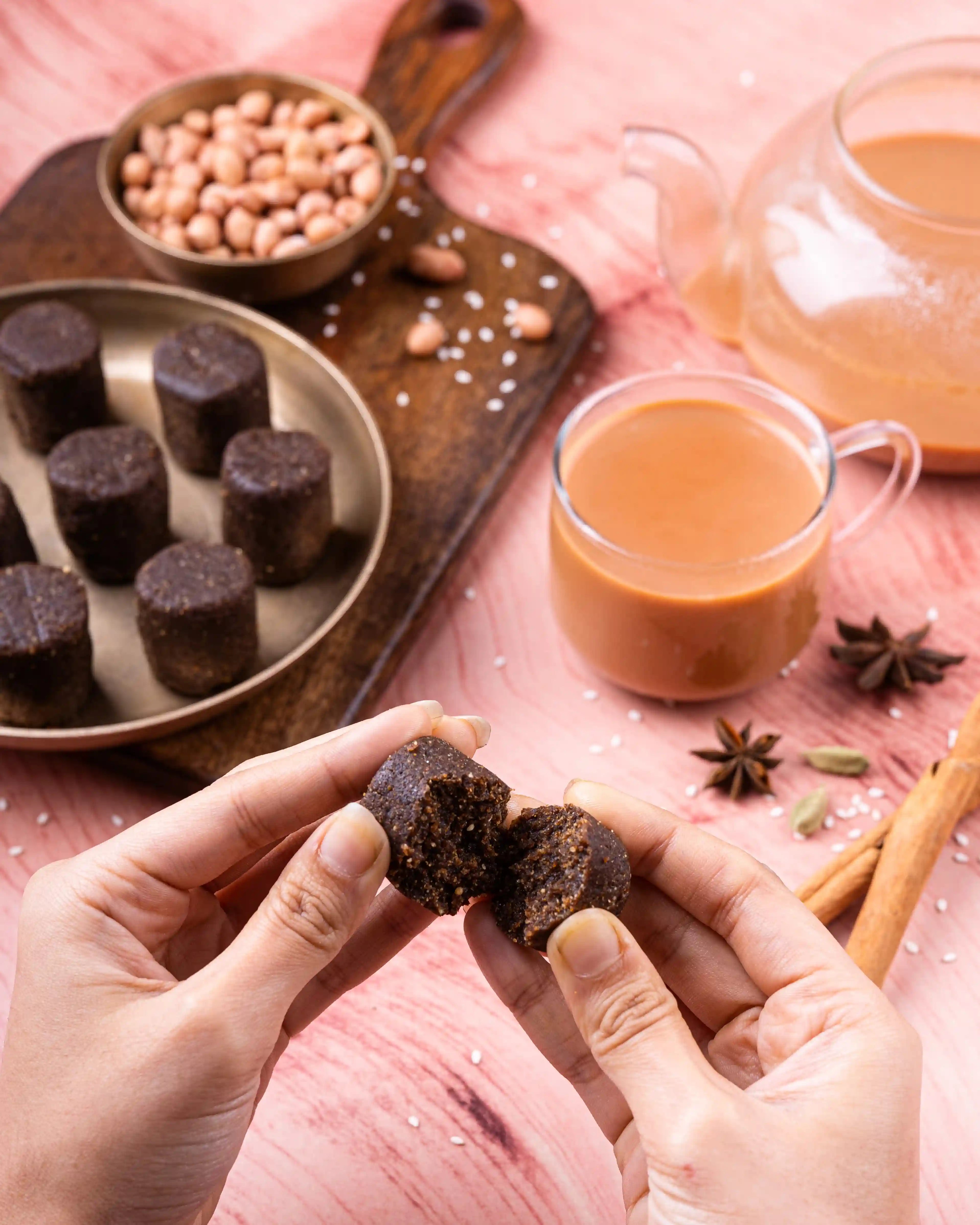 Hands breaking Aazol's Mahua Laddu (Forest Flower & Jaggery Nutri Ball) with chai